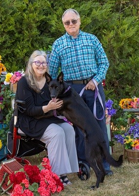 A dog with three legs, outside with a woman seated and a guy standing next to them holding a leash